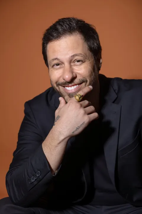 fabio kerk sorrindo, blazer escuro, mão no queixo com anel e tatuagem, fundo laranja